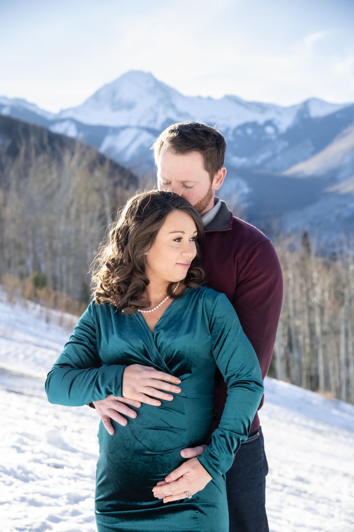 A couple shares an intimate embrace while standing in the snow with snow-capped mountains and bare trees in the background Aspen Colorado