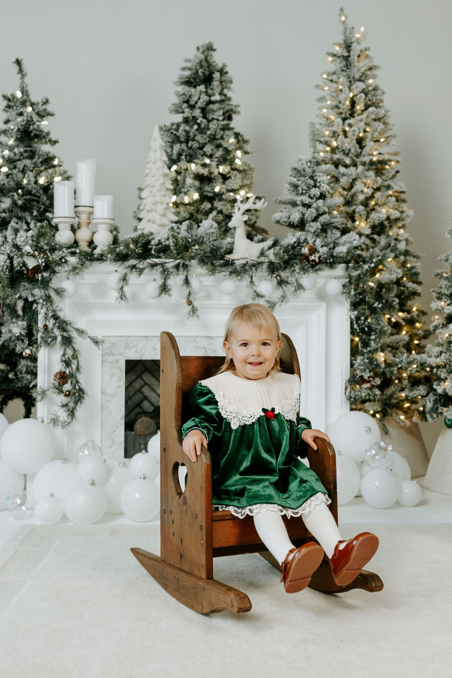 A child in emerald green velvet dress sits in a wooden sleigh against a backdrop of lit Christmas trees.