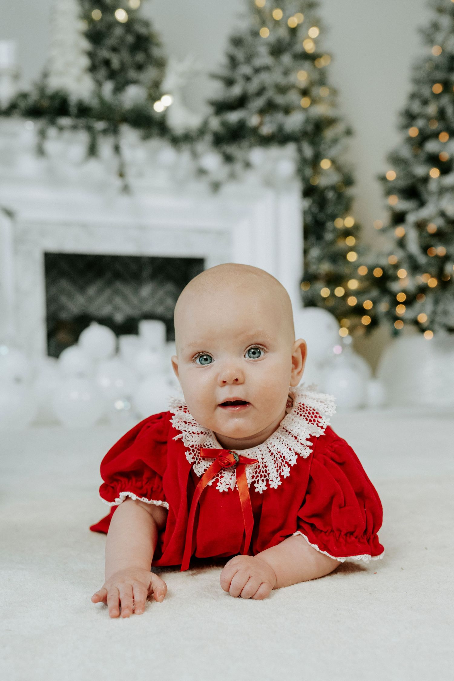 A baby in a red velvet holiday dress with white lace collar lies on a white blanket with Christmas trees in background.