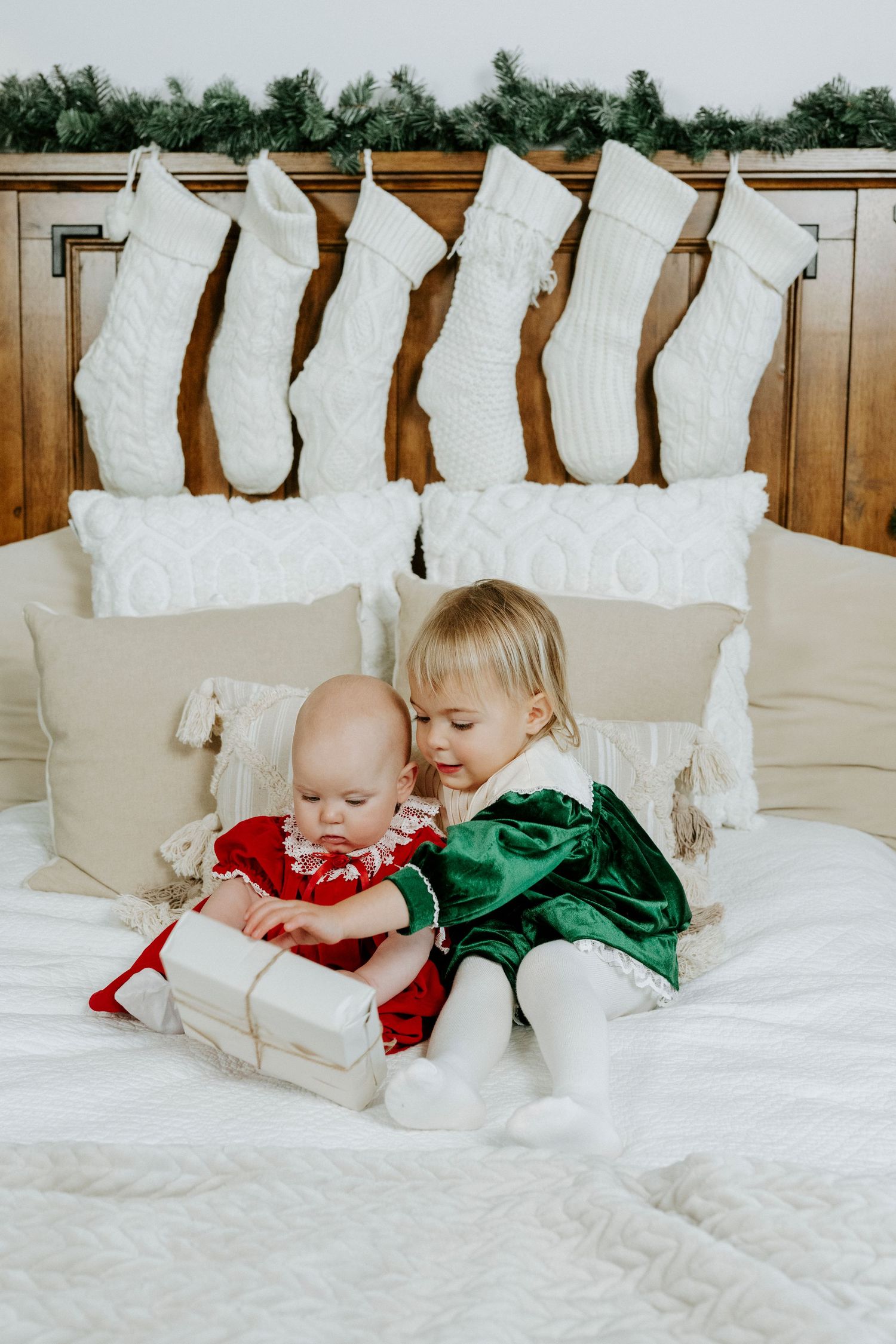 Two small children in festive red and green outfits sitting on a white bed with Christmas stockings hanging above.