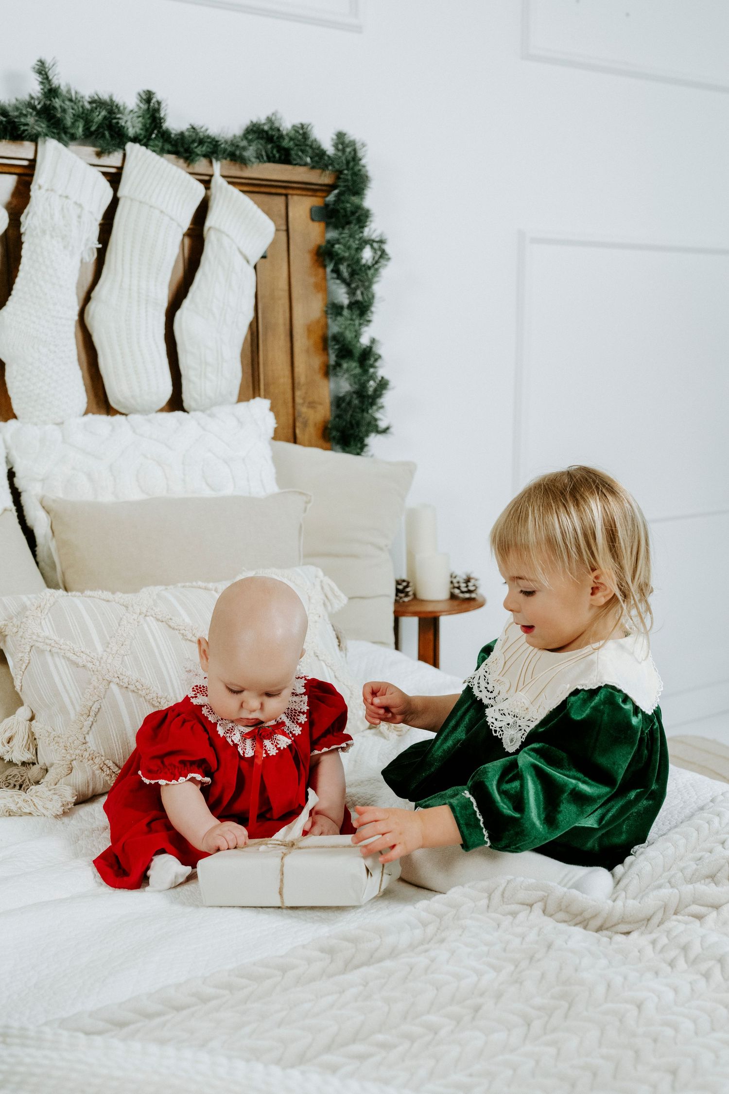 Two children in red and green holiday outfits lay on white bedding during a Christmas photoshoot.