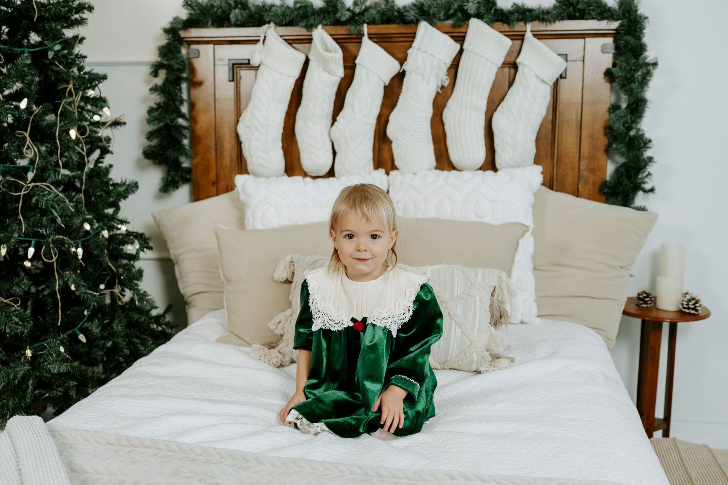 A child in green pajamas sits on a white bed with holiday decor and a Christmas tree in the background.