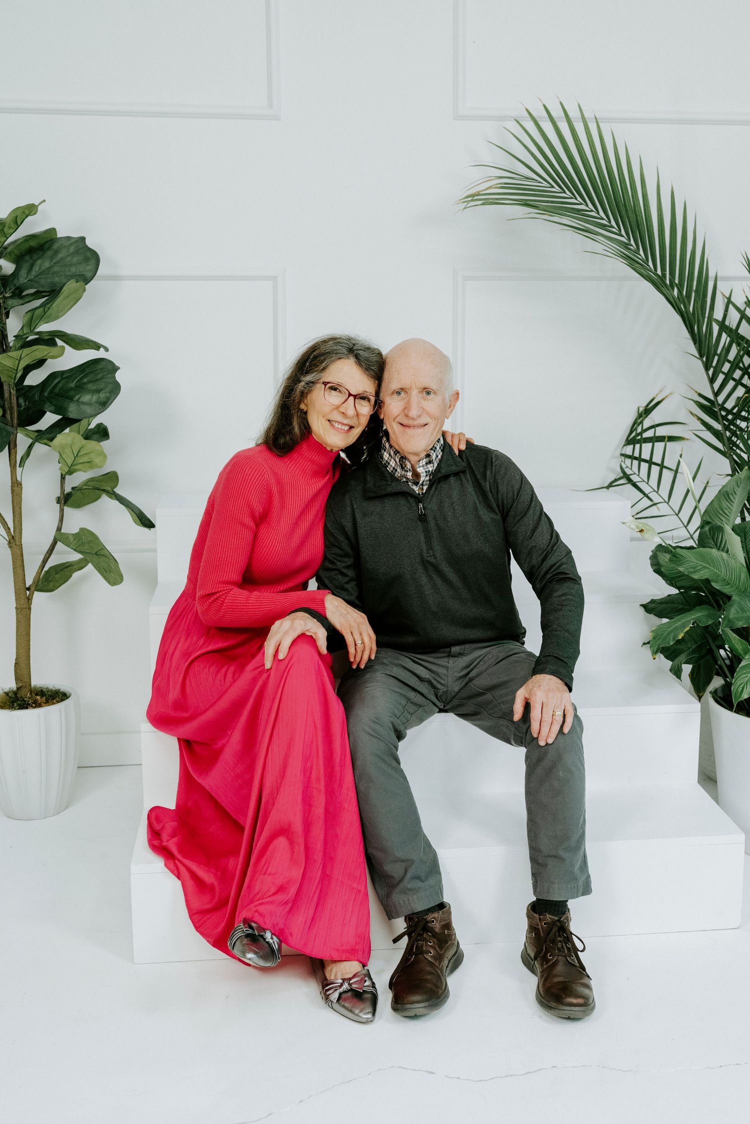 A couple in red and black attire pose together on white steps with tropical plants in the background.