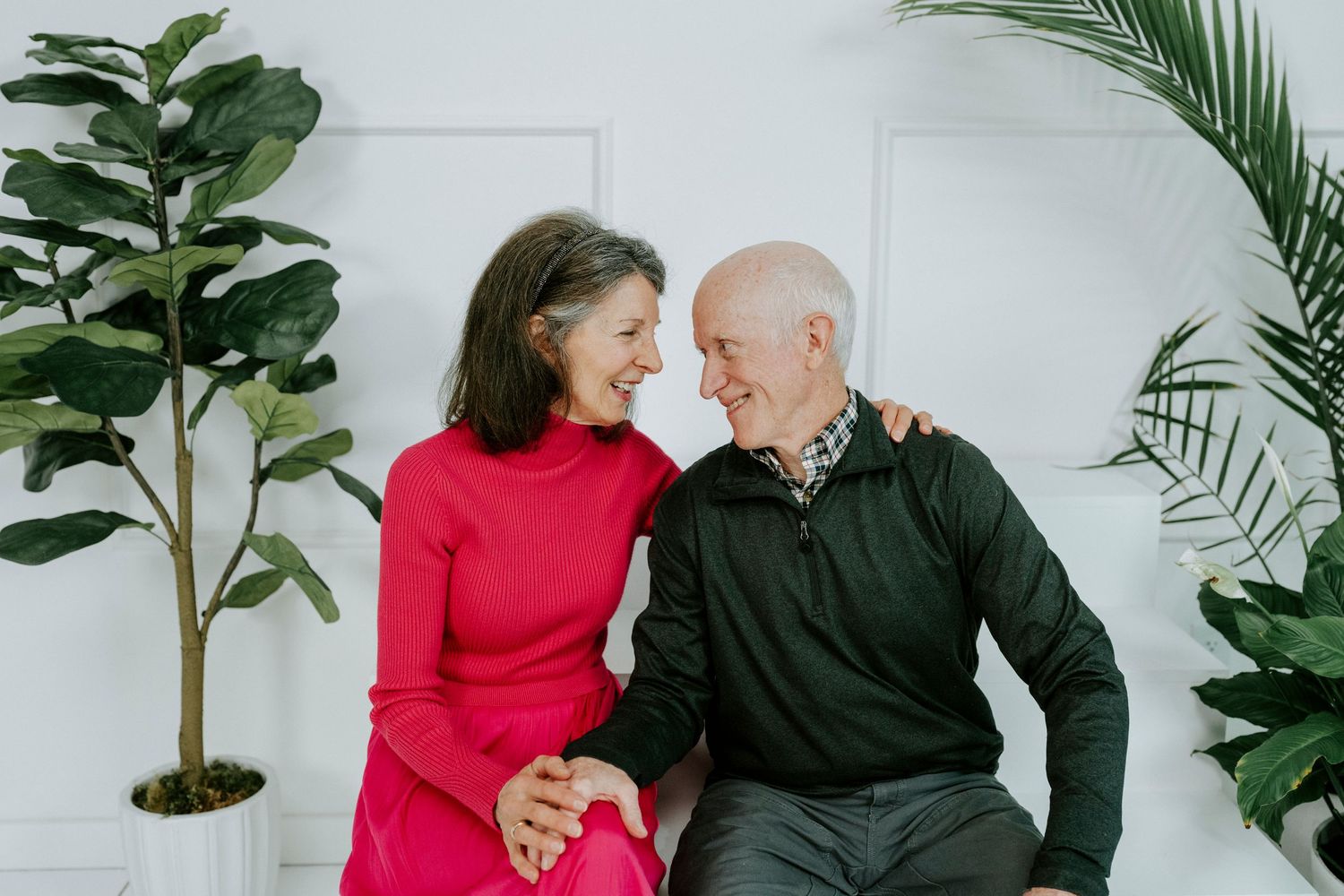 Two people share a joyful moment while sitting together against a white wall with green plants in the background.