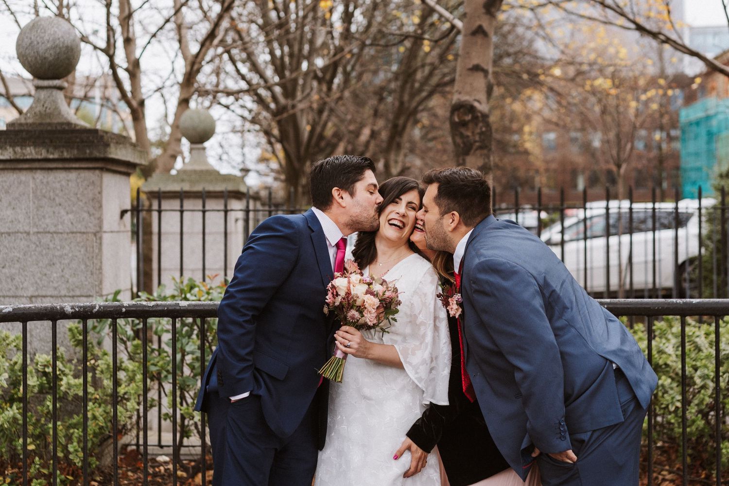 Three people in formal attire share a sweet moment by an iron fence while holding a pink floral bouquet.