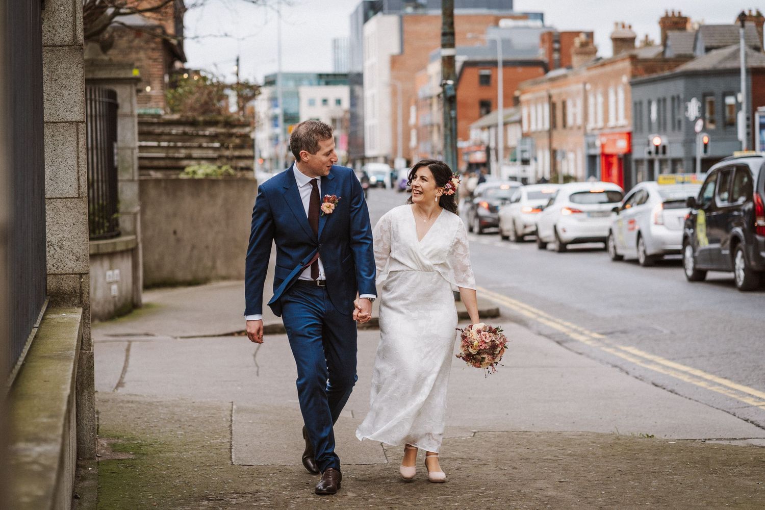 A couple in wedding attire walks down an urban street lined with parked cars on a cloudy day.