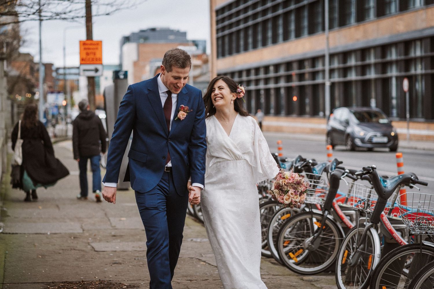 Newlyweds walk together on a city sidewalk with bicycles parked in the background.