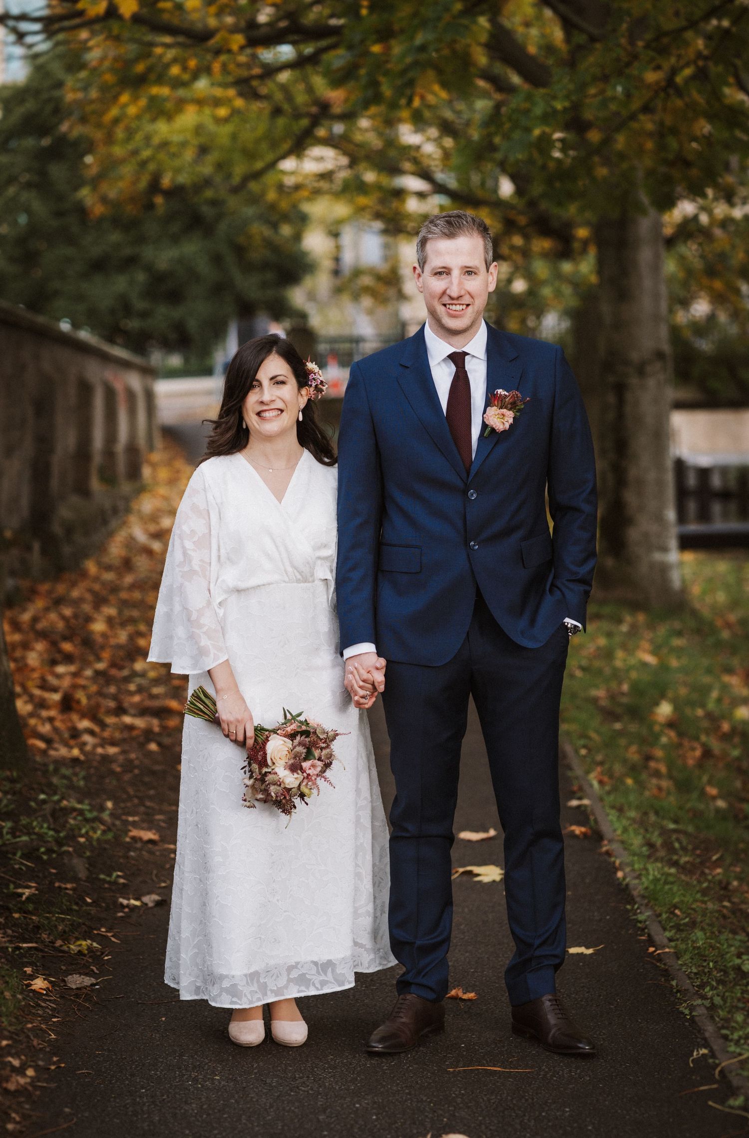 A couple in wedding attire walks along a tree-lined autumn path with fallen leaves on the ground.