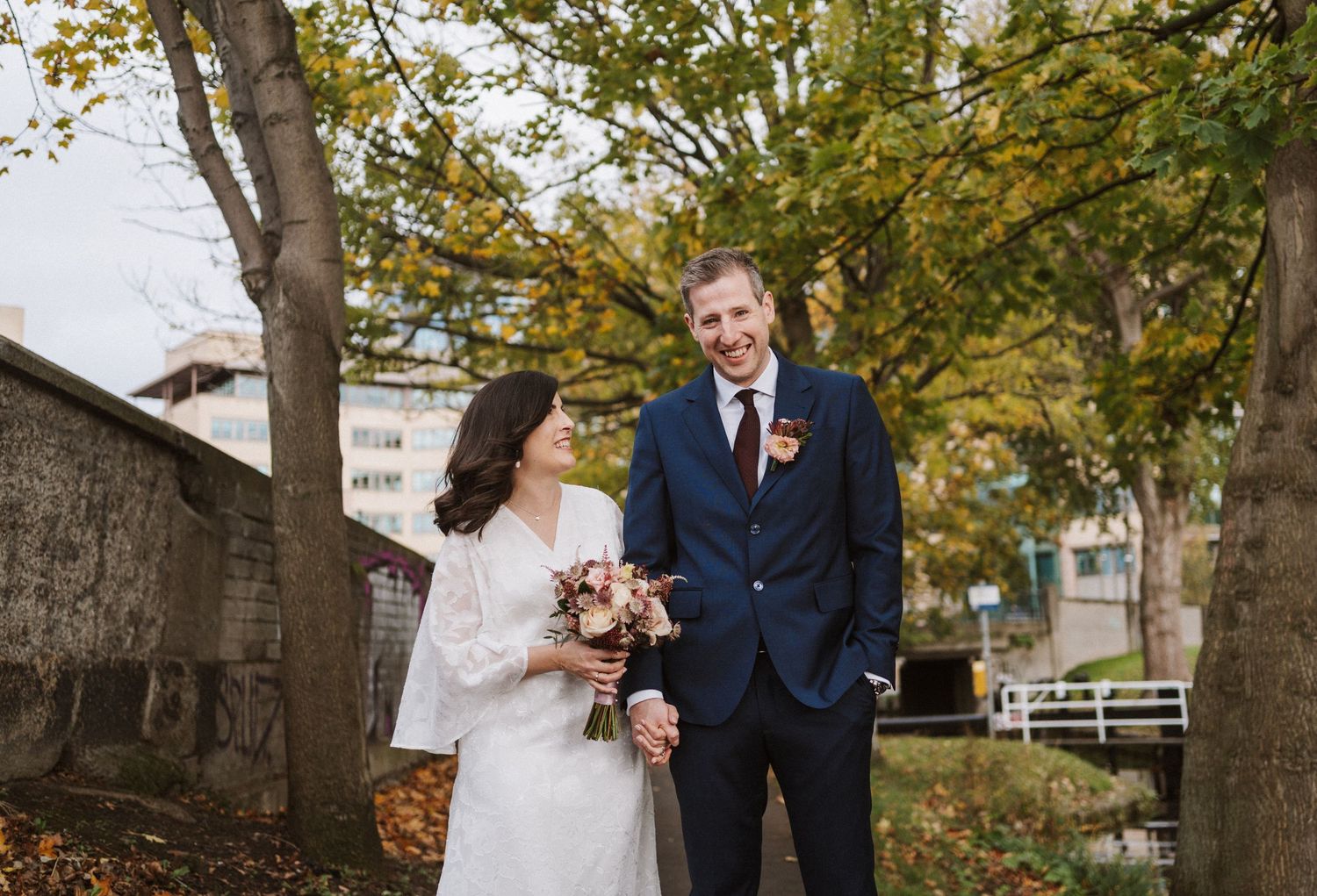 A couple shares a joyful moment on their autumn wedding day surrounded by golden leaves and trees.