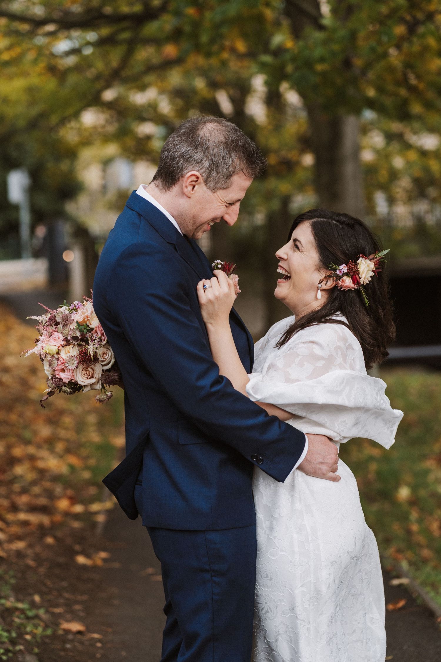 A couple in wedding attire shares joyful laughter together during an autumn photoshoot with fallen leaves in the background.