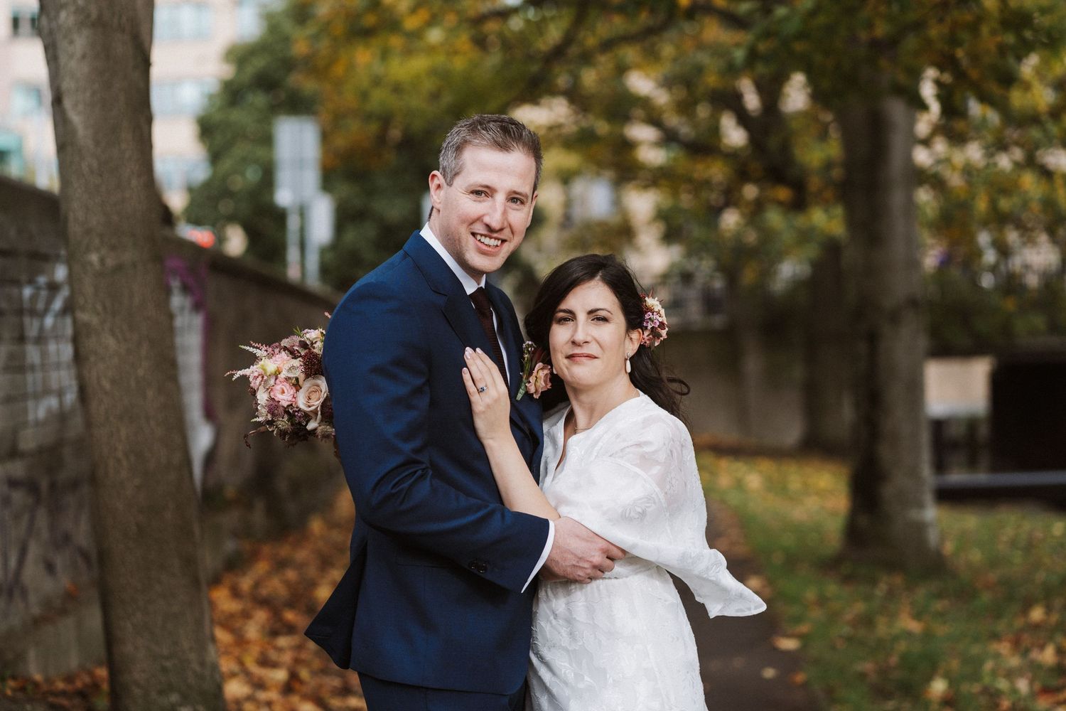 A couple in wedding attire embraces and smiles while standing among autumn trees in a park setting.