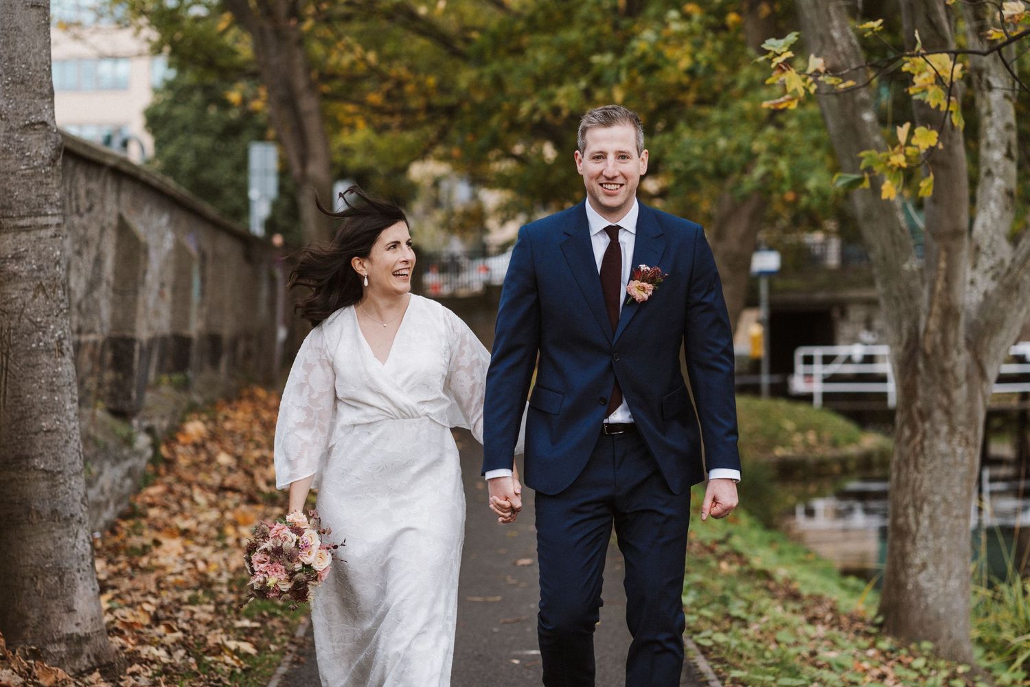 A couple in wedding attire walks together through a park with autumn leaves on the ground.