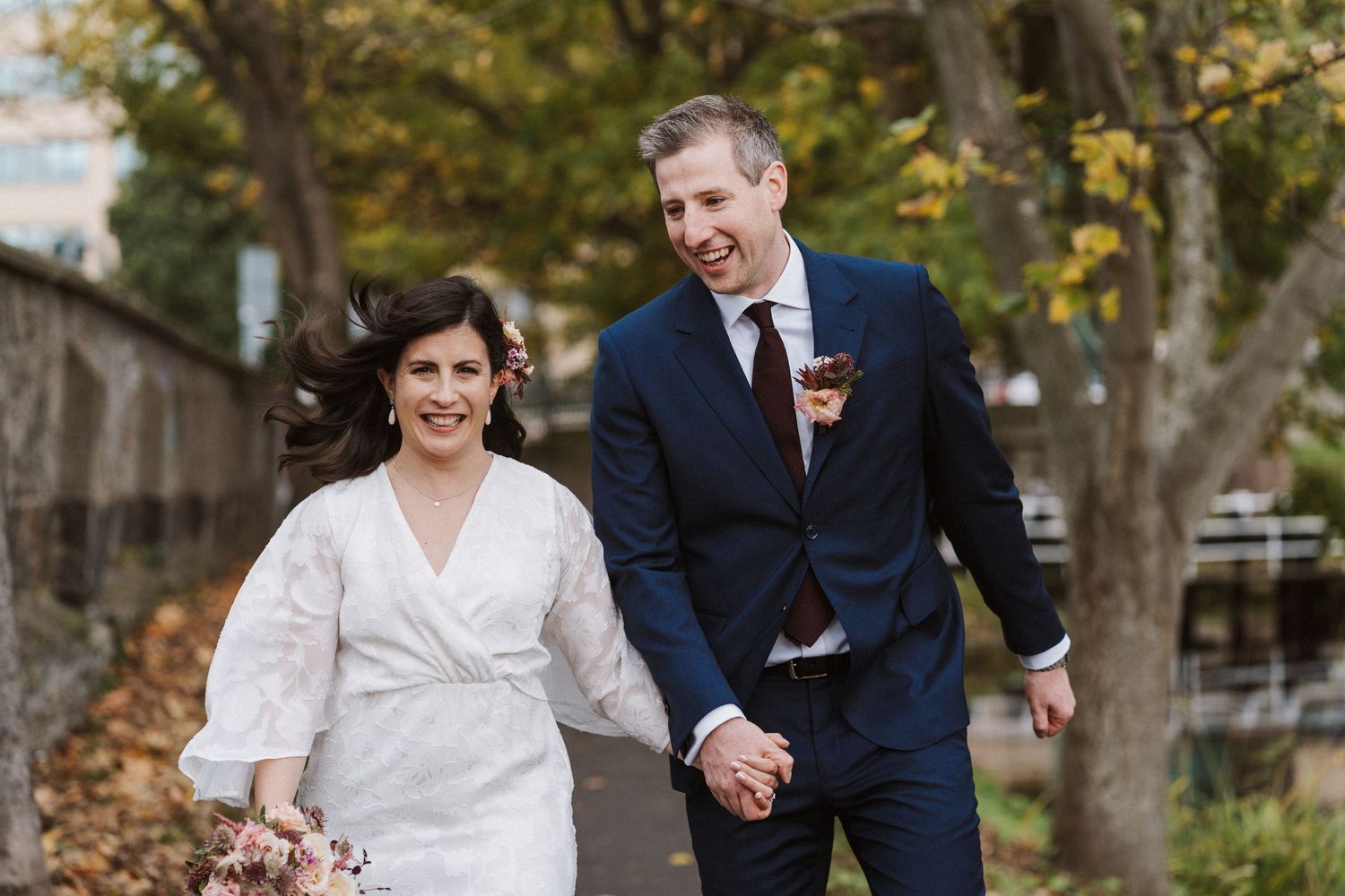 A happy couple walks hand in hand through a wooded park with autumn foliage in the background.