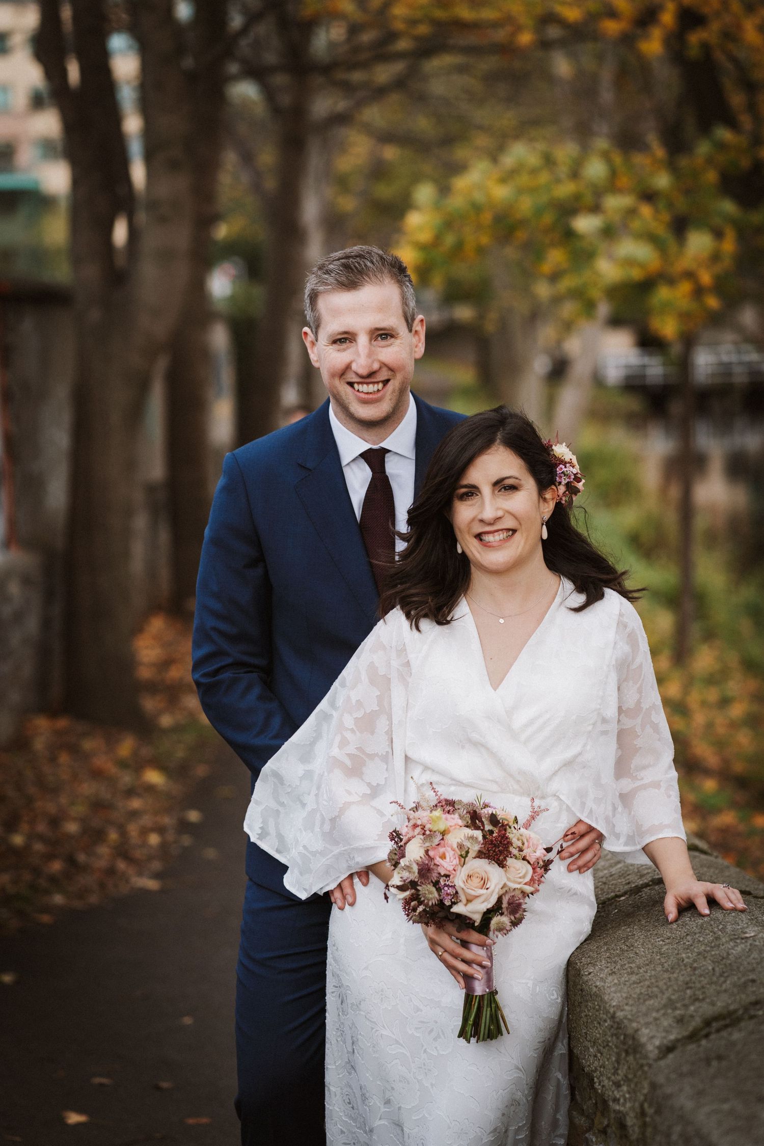 A couple poses for wedding photos in a park with fall trees, the bride holding a pink and white bouquet.