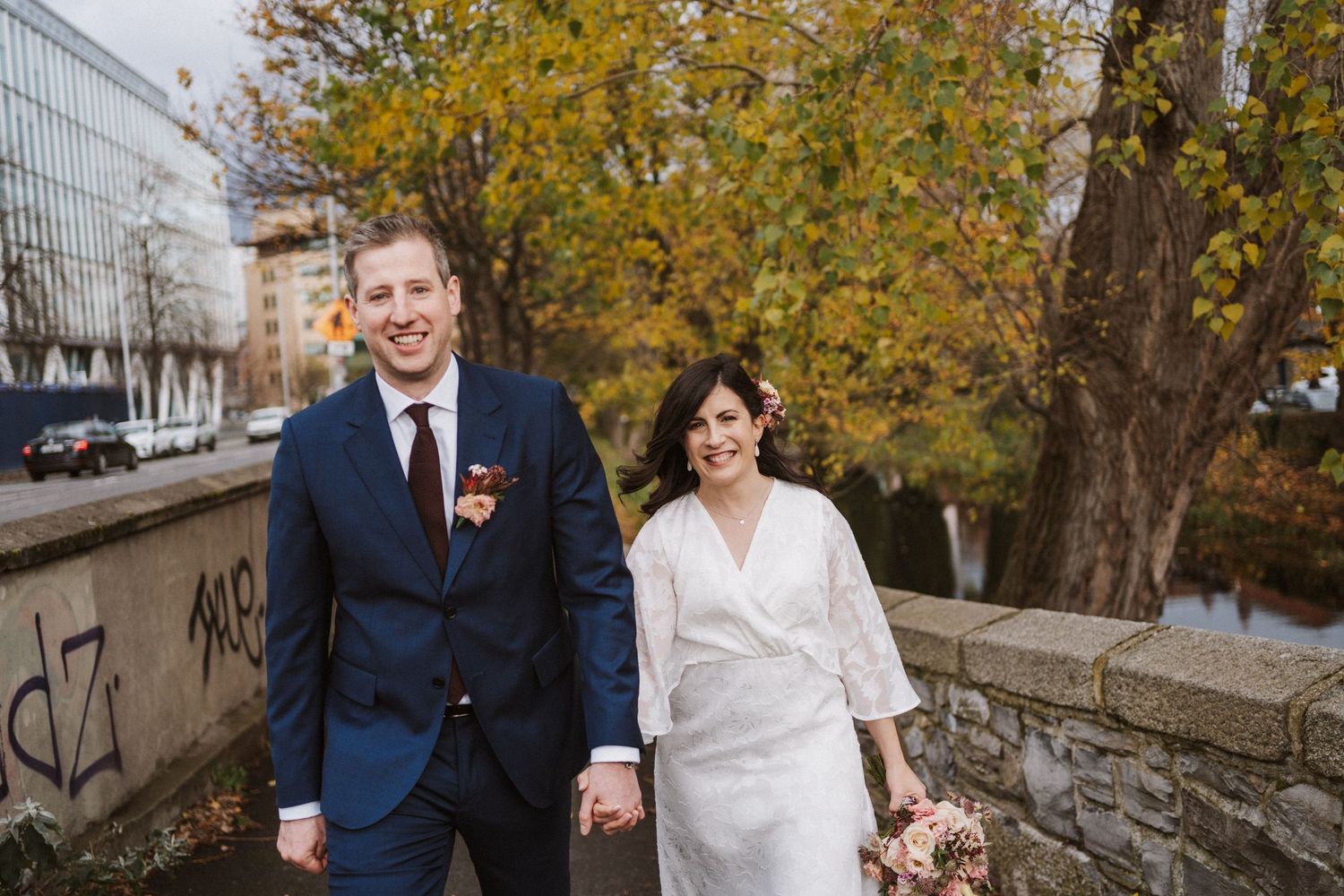 A couple in wedding attire walk together through a scenic autumn park with golden leaves on a sunny day.