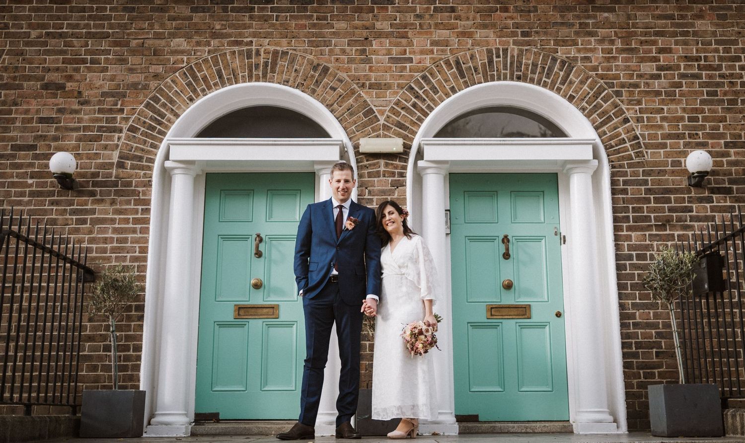 A couple stands between two matching turquoise doors on a classic brick building with arched doorways.