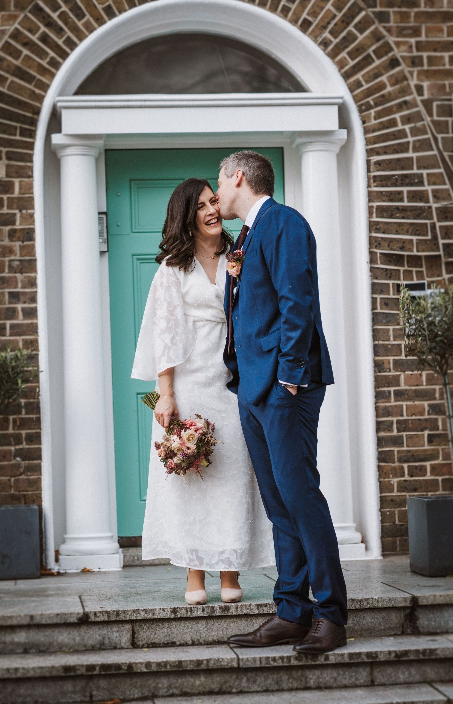 A couple shares a tender moment in front of a turquoise door in an arched brick entryway.