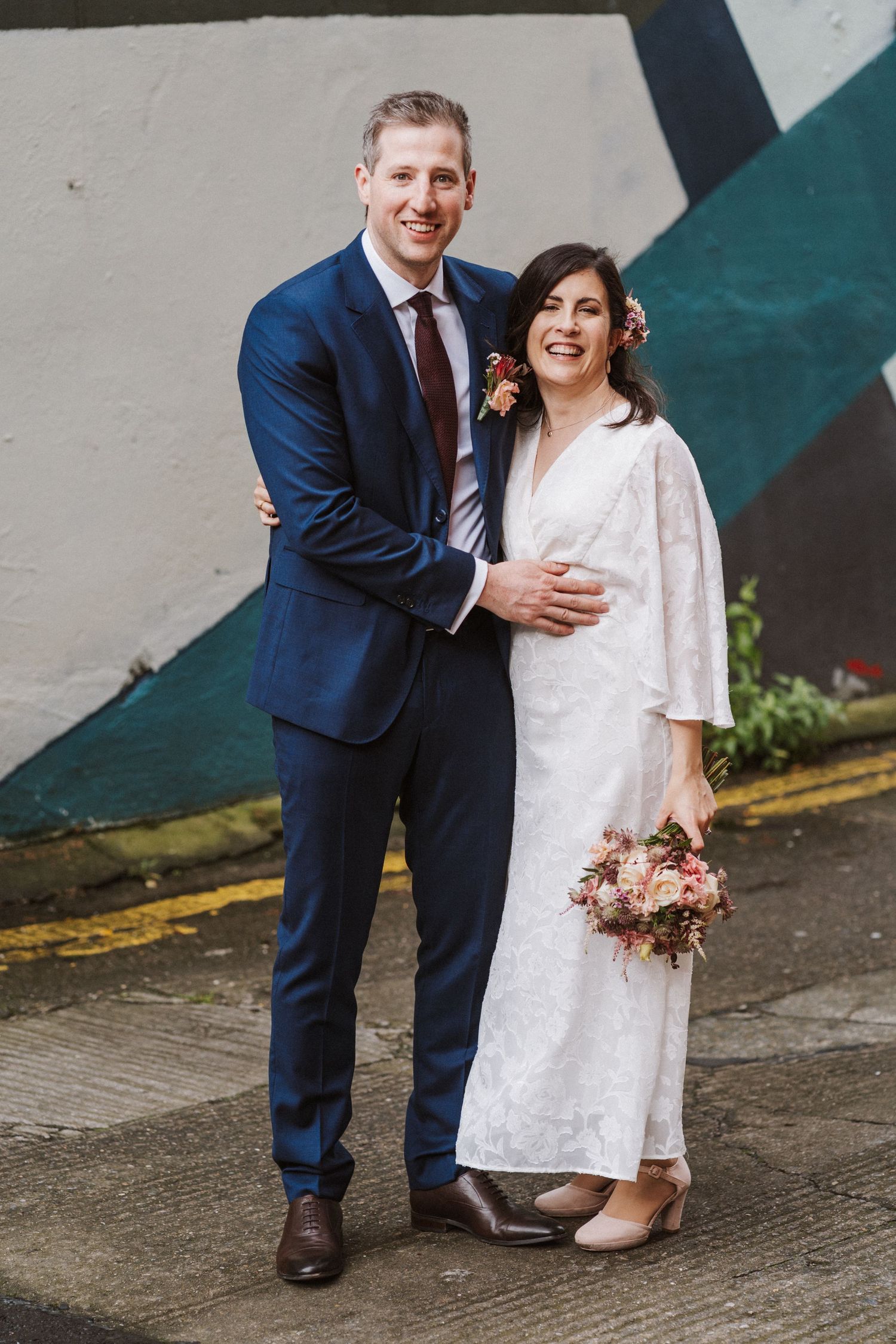 A couple in wedding attire shares a joyful moment against a wall with colorful geometric designs.