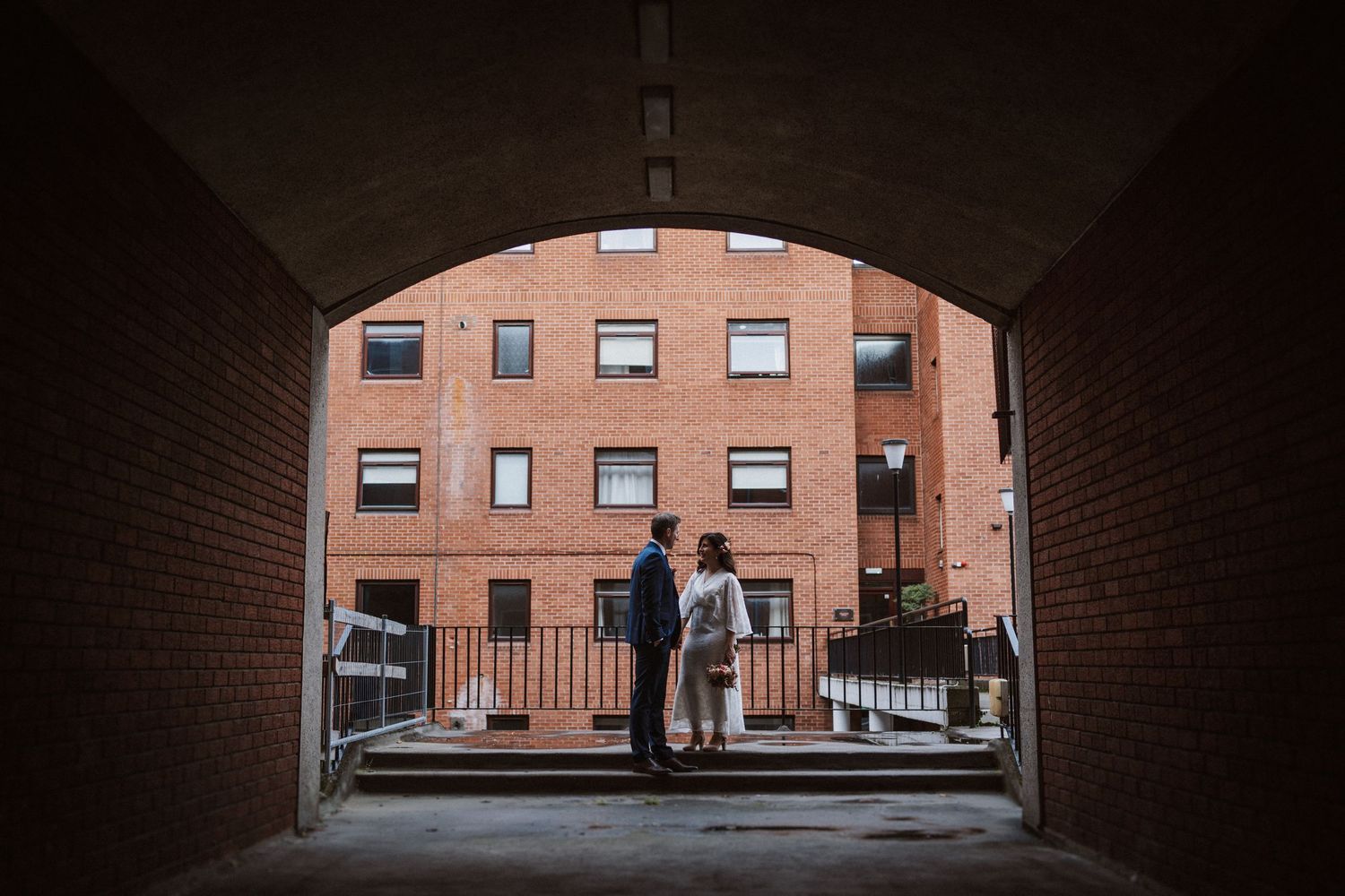 A dark brick tunnel or passageway leading to a bright courtyard with red brick buildings visible at the end.