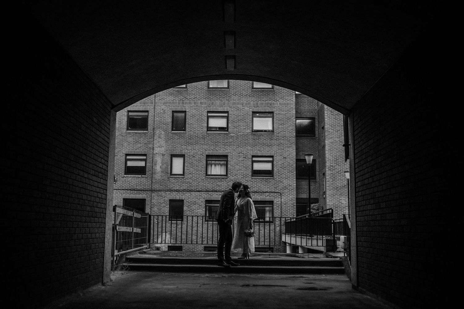 A silhouetted figure stands in a dark tunnel archway overlooking a brick apartment building in black and white.