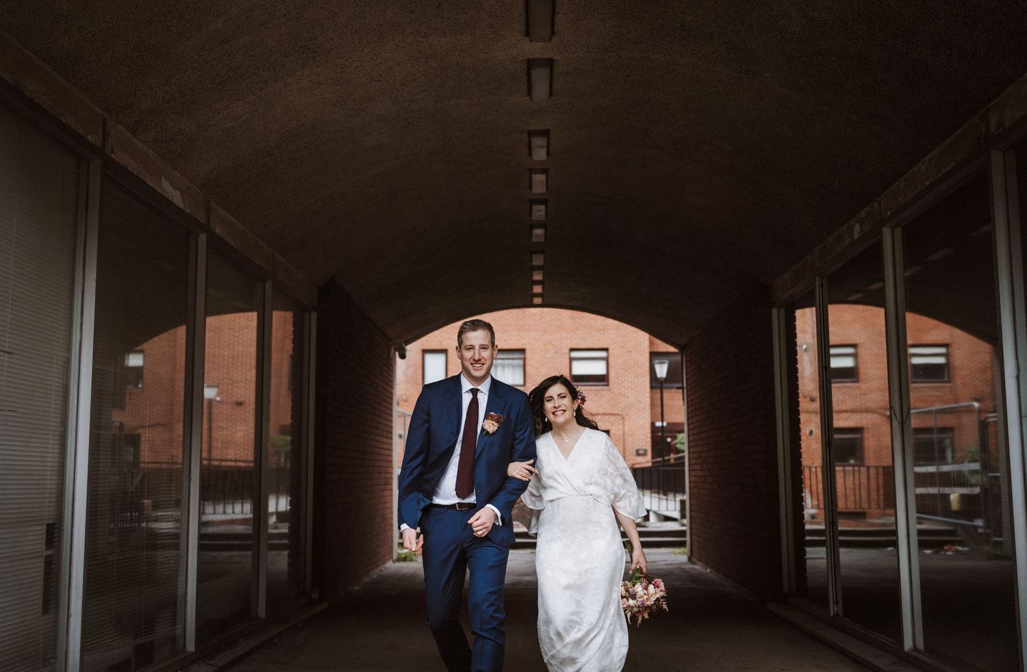 A panoramic wedding photo showing a couple in formal attire standing in a long industrial-style covered walkway.