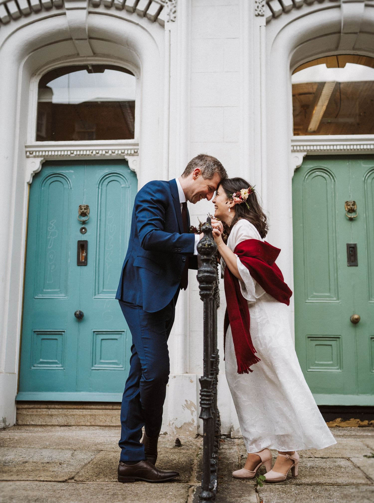 A romantic couple poses between two turquoise doors on a white building exterior with ornate architectural details.