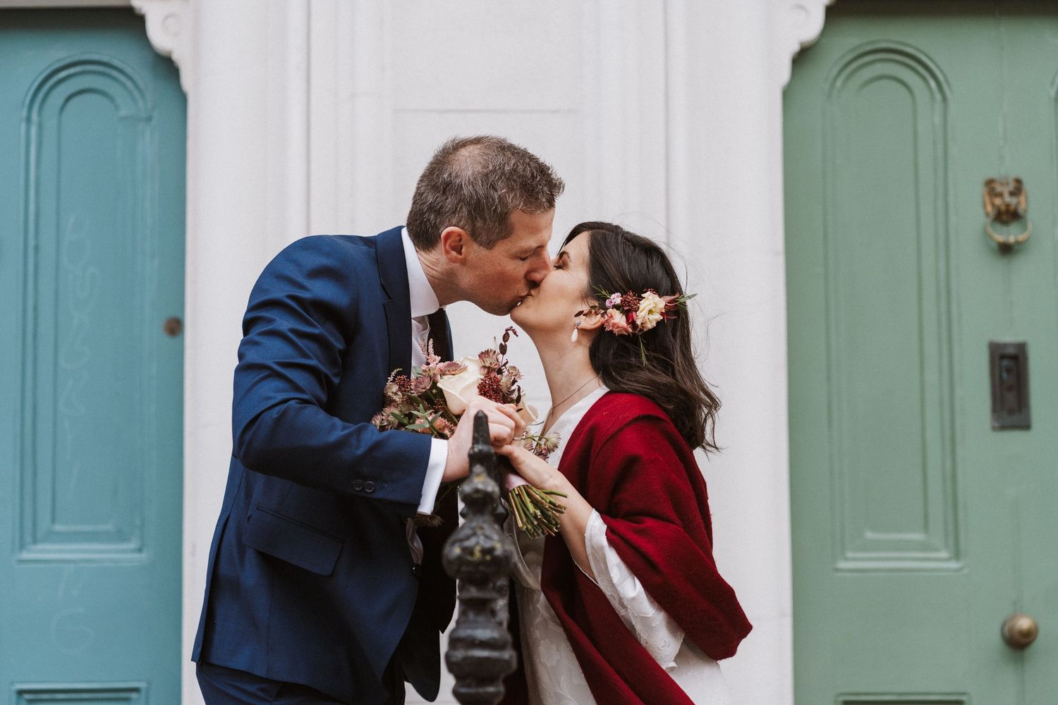 A couple shares a romantic kiss in front of a row of pastel-colored doors.