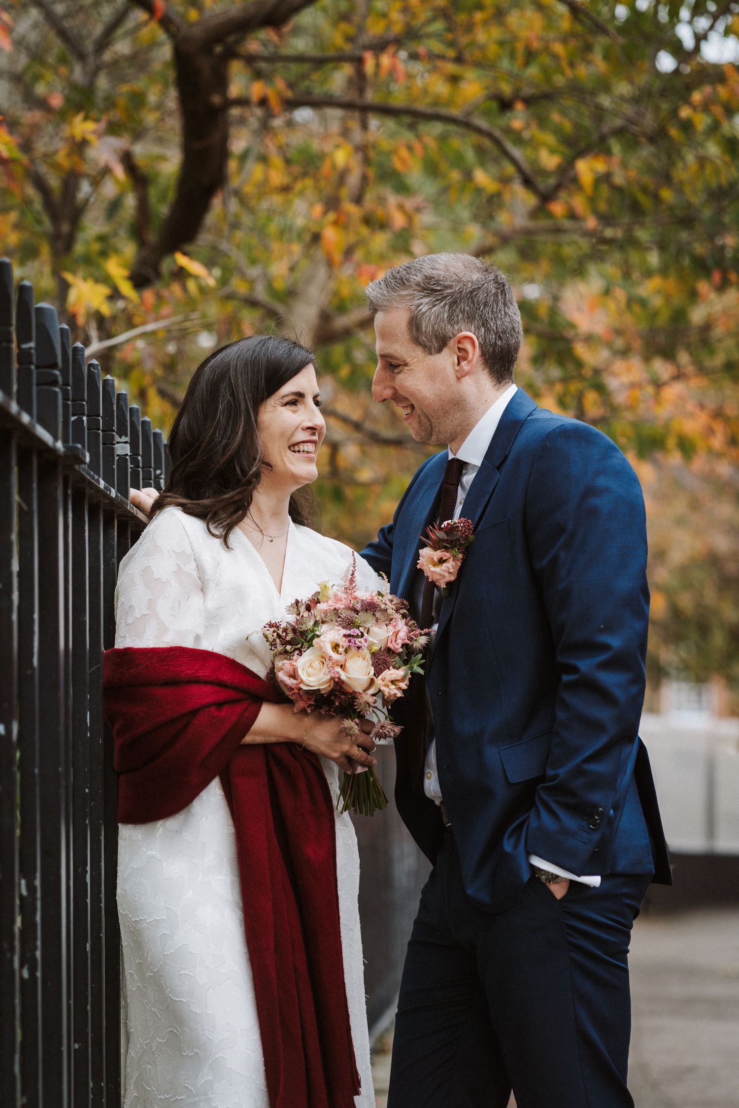 A couple shares joyful moments in their wedding attire against a backdrop of autumn foliage and iron railings.