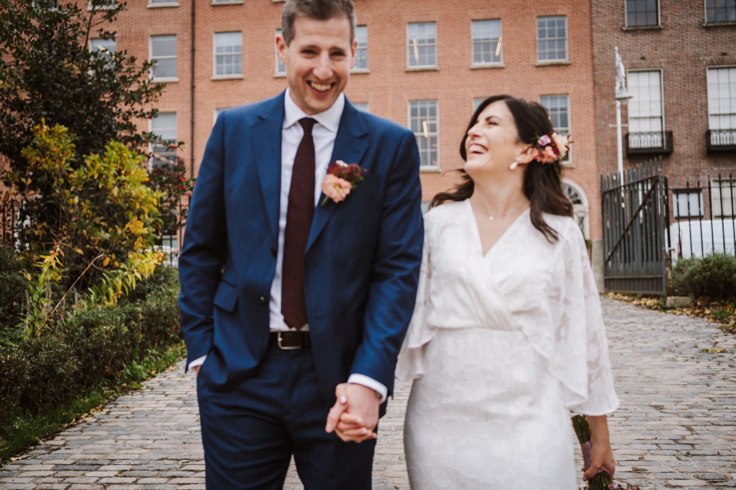A couple shares a joyful moment on their wedding day, wearing a navy suit and white dress on a stone path.