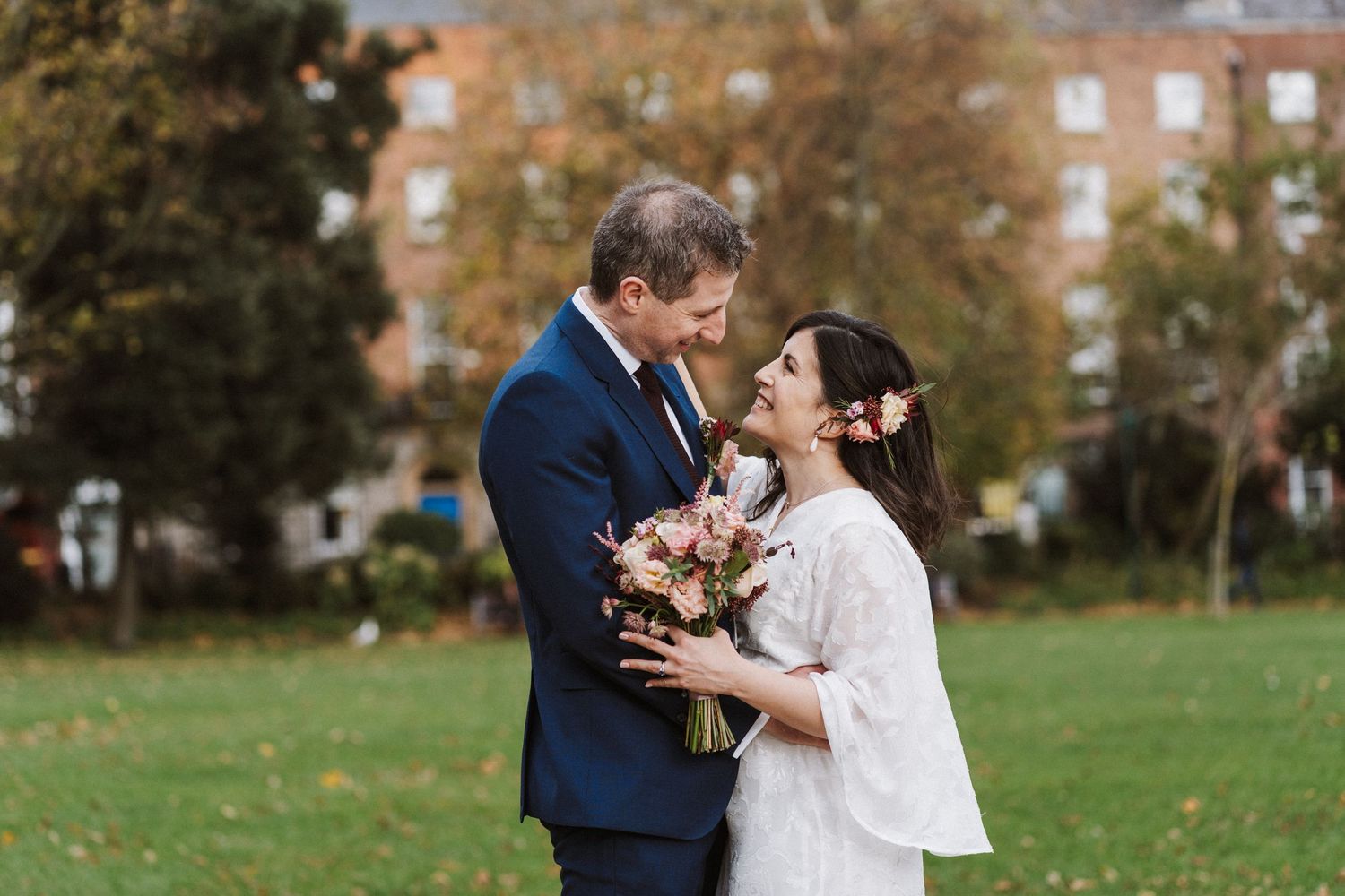 A romantic wedding couple embraces outdoors in a park during autumn with golden fall foliage in the background.