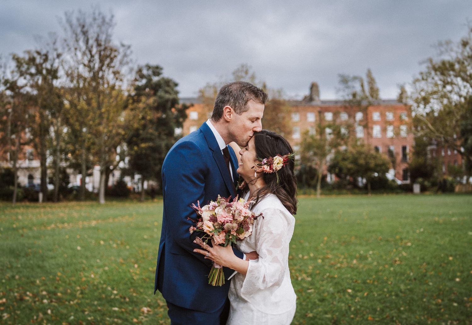 A couple shares a romantic moment outdoors on a fall day with brick buildings in the background.