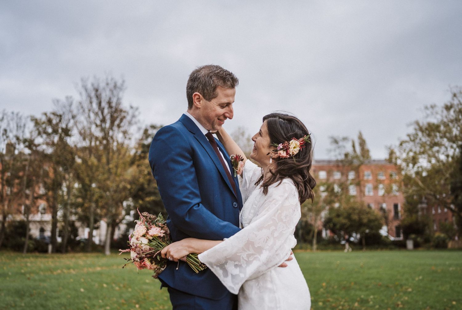 A couple embraces in a tender moment on a grassy lawn with autumn trees and historic buildings behind them.