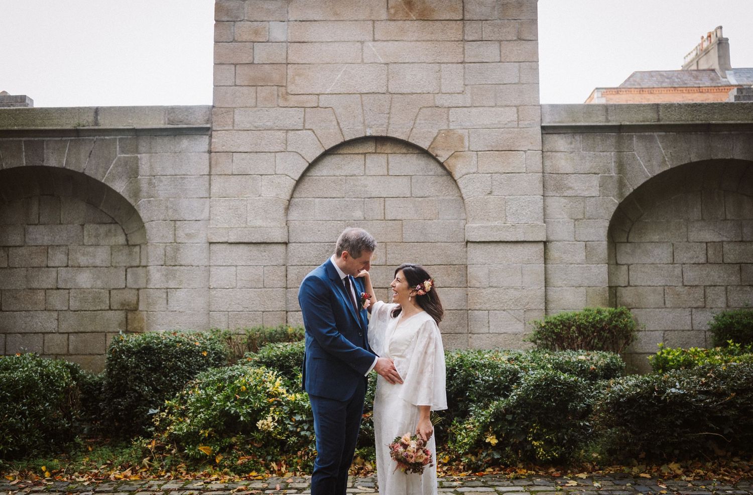 A romantic wedding couple shares intimate moments under stone archways and against garden walls.