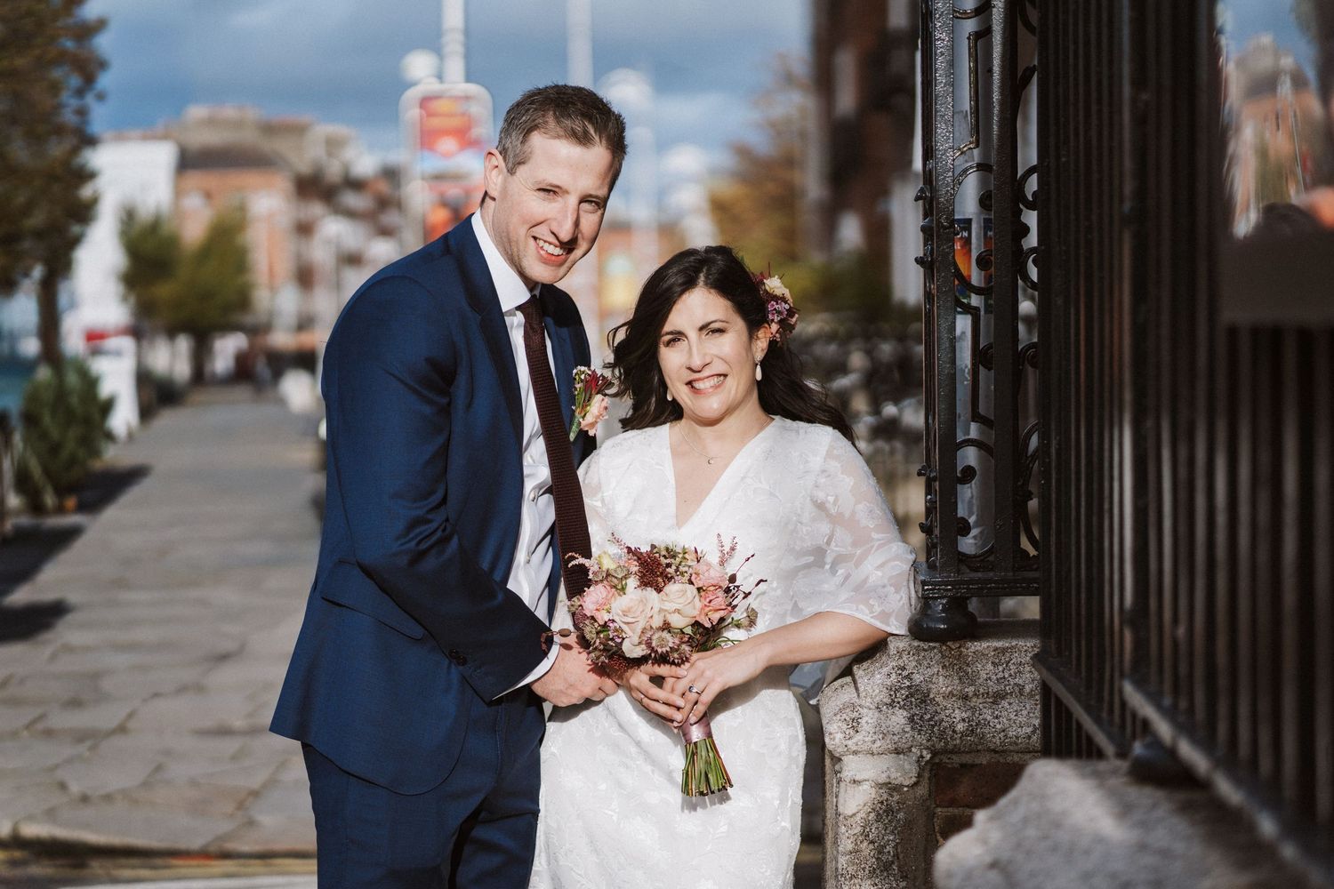 A wedding couple poses together on a cobblestone street with brick buildings and black iron fences in the background.