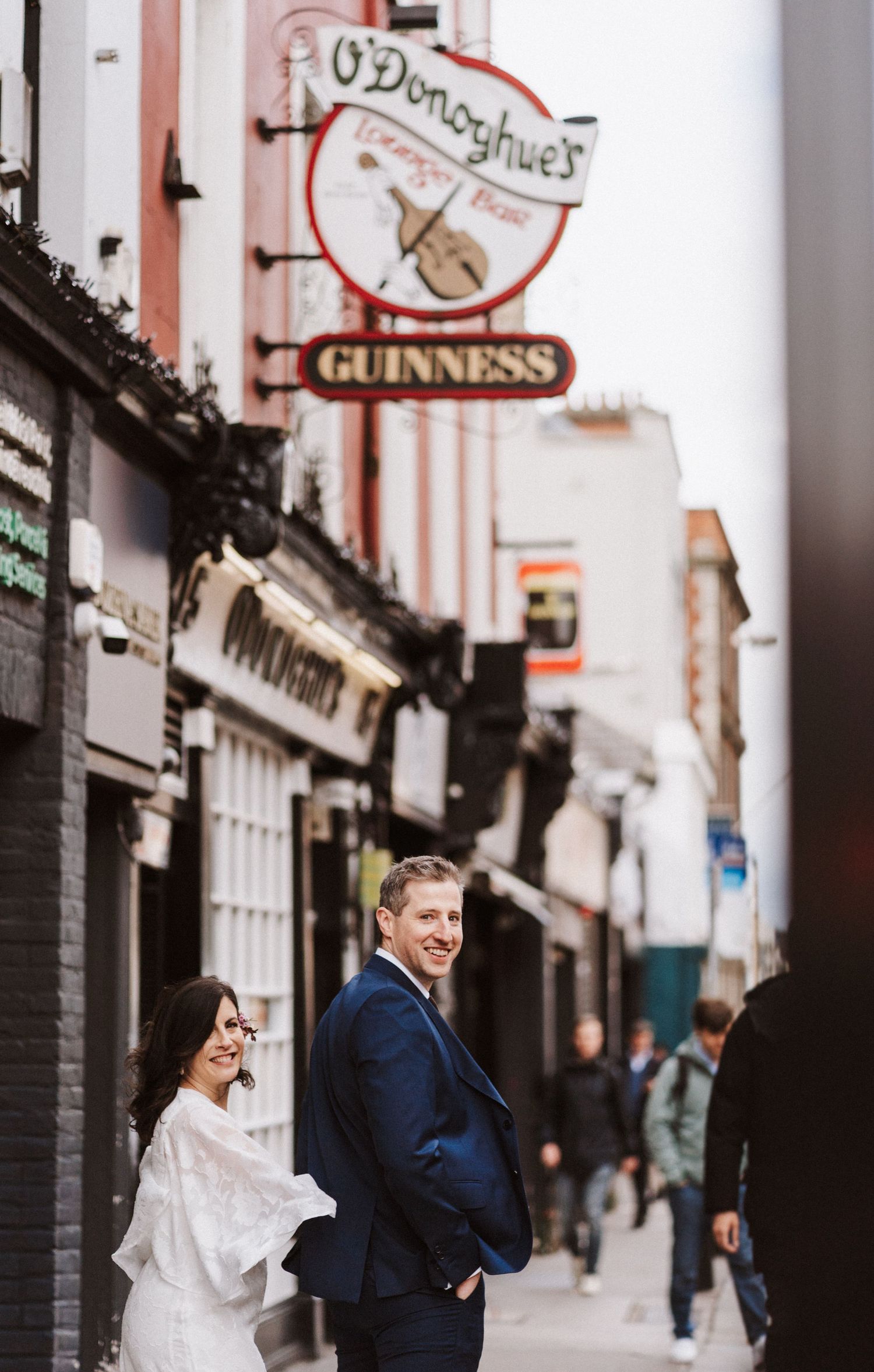 A couple in wedding attire walks down a charming European street lined with shops and Guinness signs.