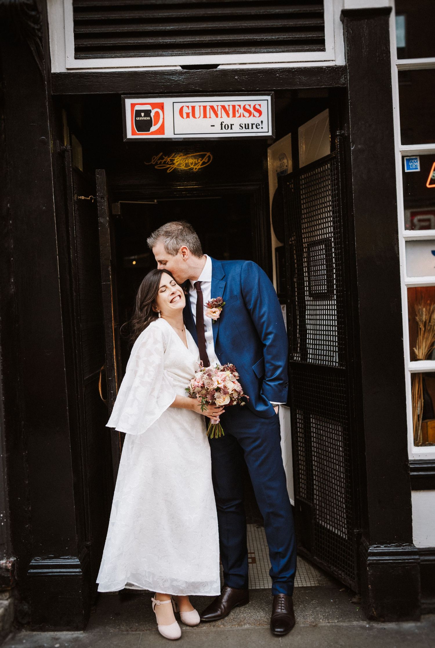 A couple shares an intimate moment outside a pub with a Guinness sign, the bride holding a pink bouquet.