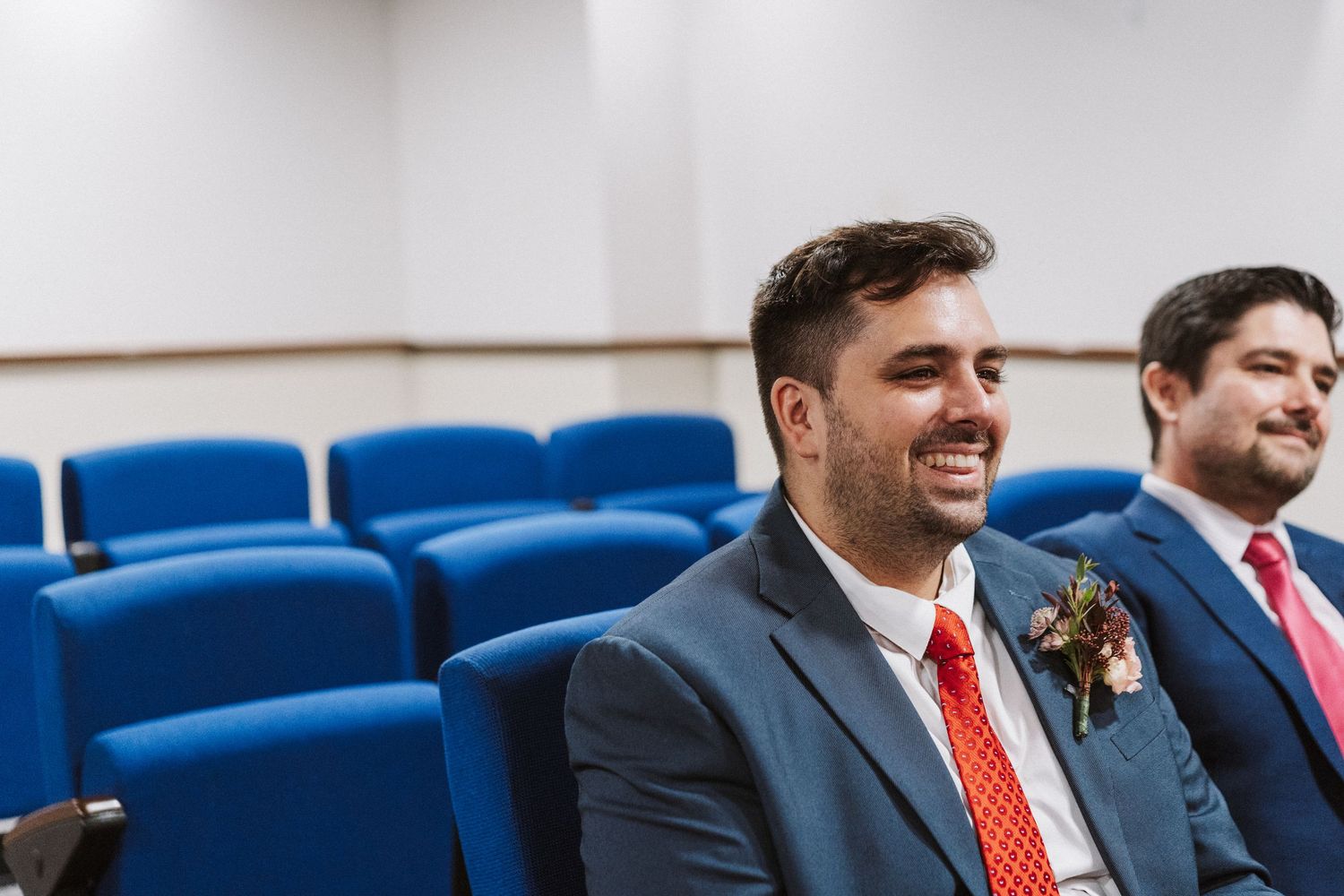 Business professionals in suits and red ties sit in a row of blue theater seats during a corporate event.