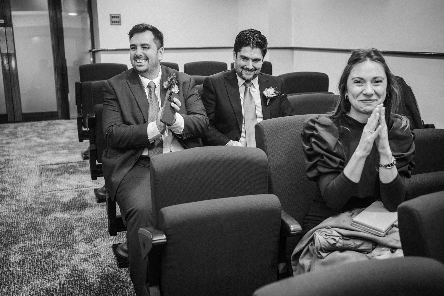A group of people sitting in a row on black chairs laugh and smile during a casual gathering.