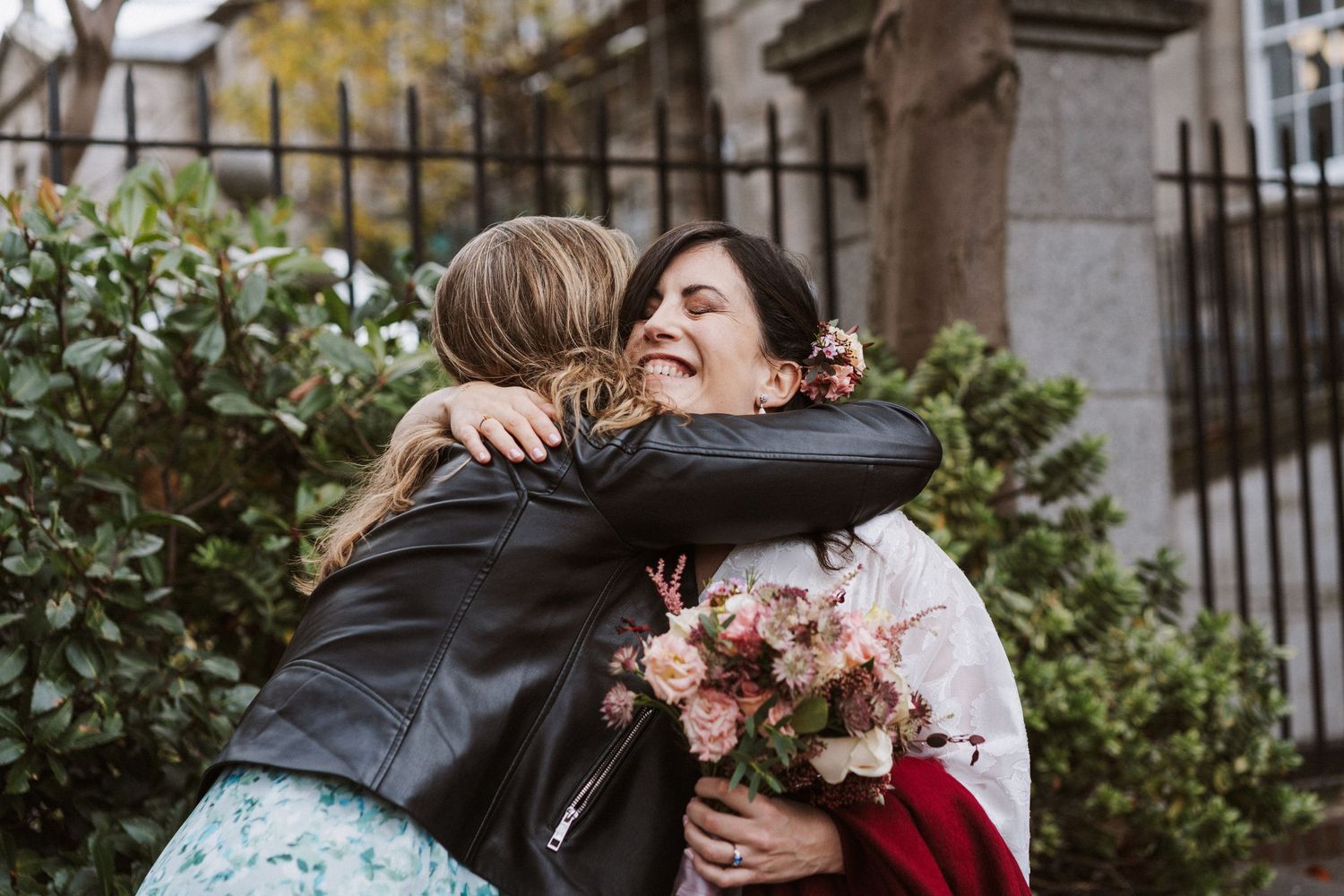 A couple shares a tender embrace outdoors in front of a stone building with autumn foliage in the background.