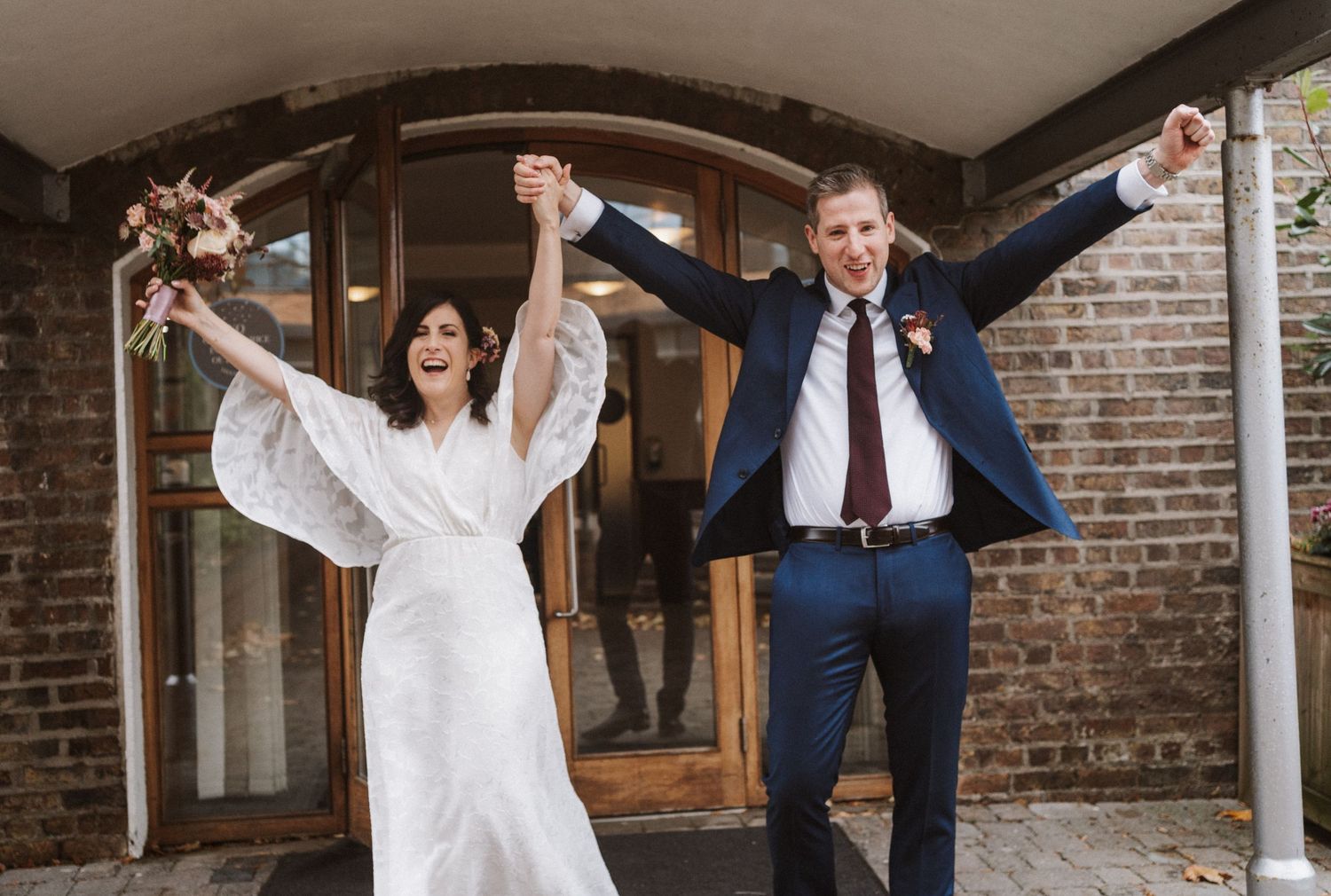 Newlywed couple celebrates with raised arms in joyful moment outside brick church entrance after wedding ceremony.