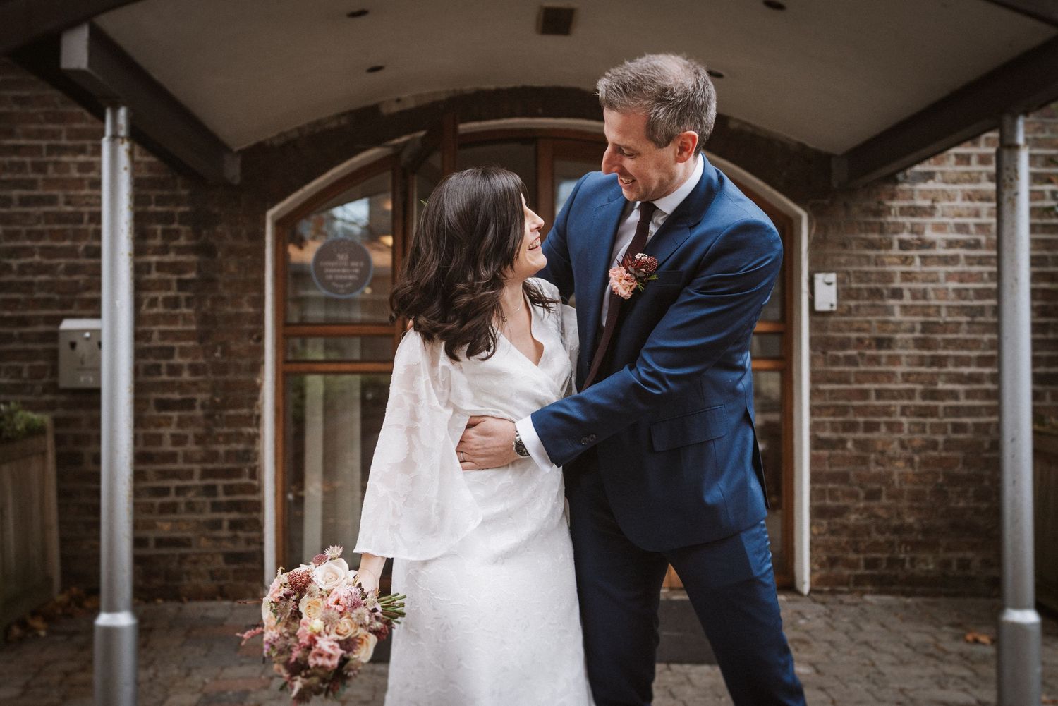 A bride and groom share a romantic moment together outside a brick building with a pink floral bouquet.