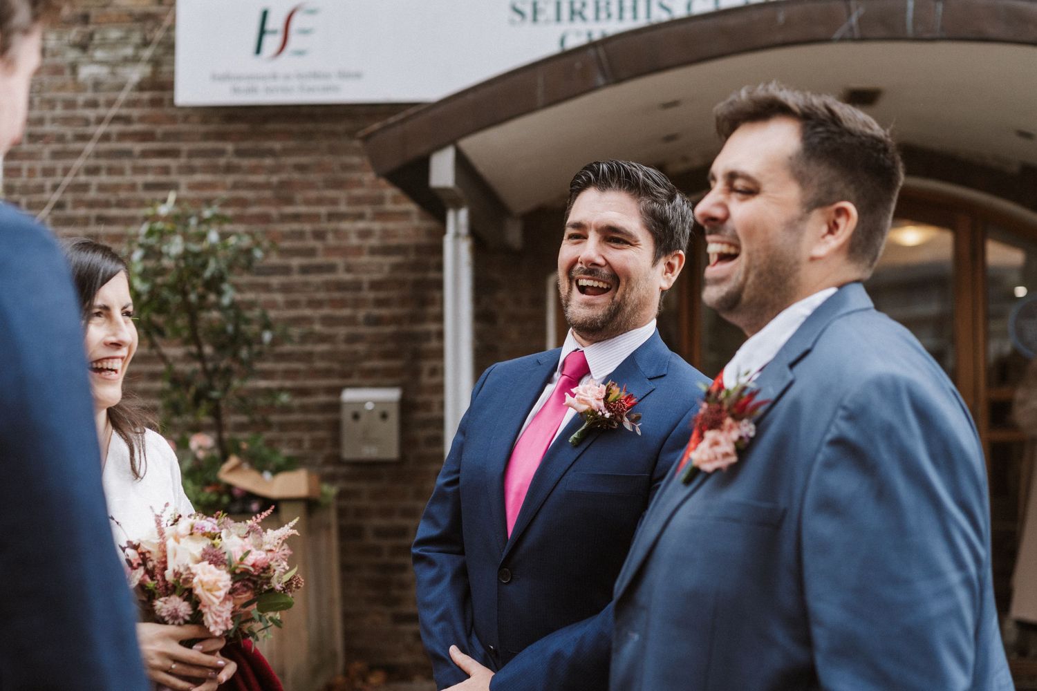 Joyful wedding party laughs together while standing outside a brick building during a celebration.