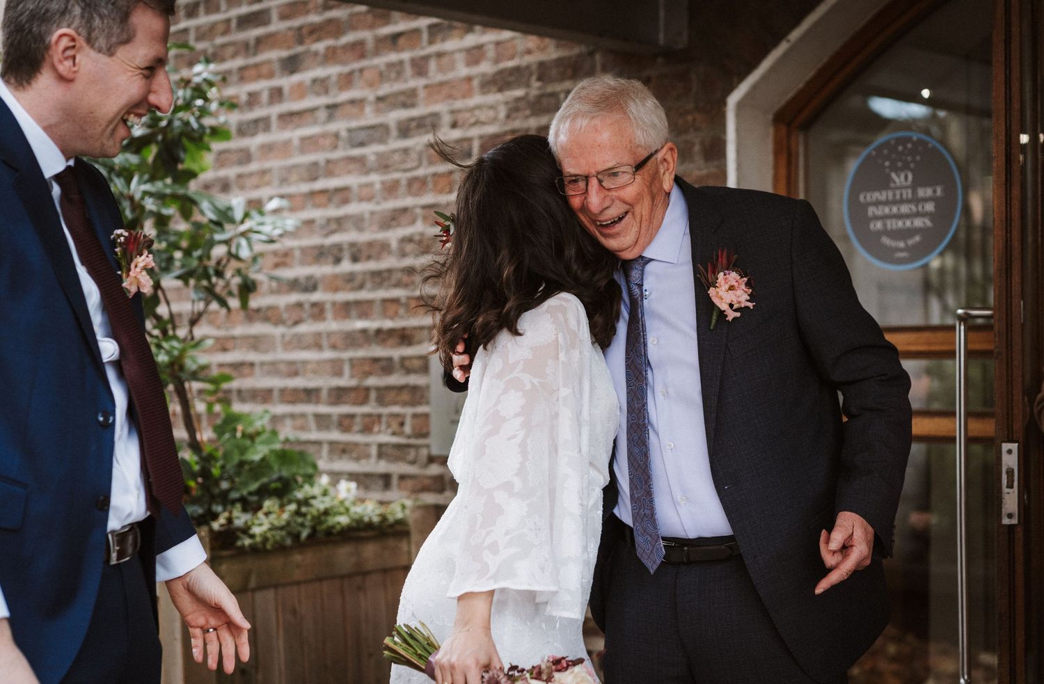 A joyful moment shared between guests outside a rustic brick building entrance with a decorative boutonniere.