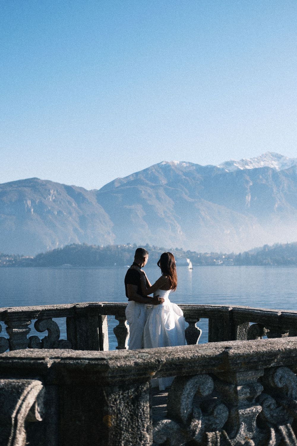 Silhouettes of people enjoying scenic views of Lake Como with snow-capped mountains in the background on a clear day.