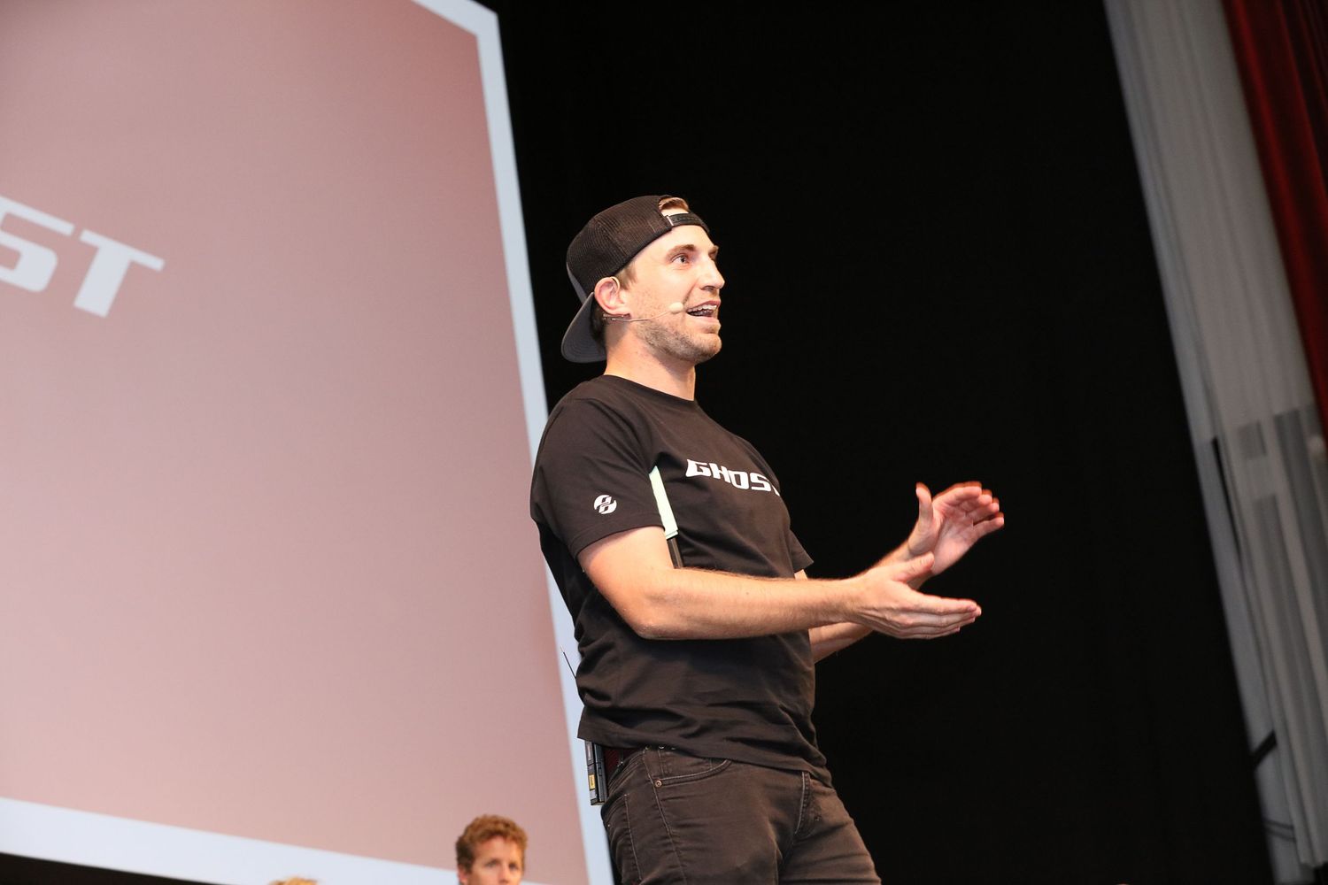 A speaker in a black t-shirt gestures while presenting on stage at a conference or event.