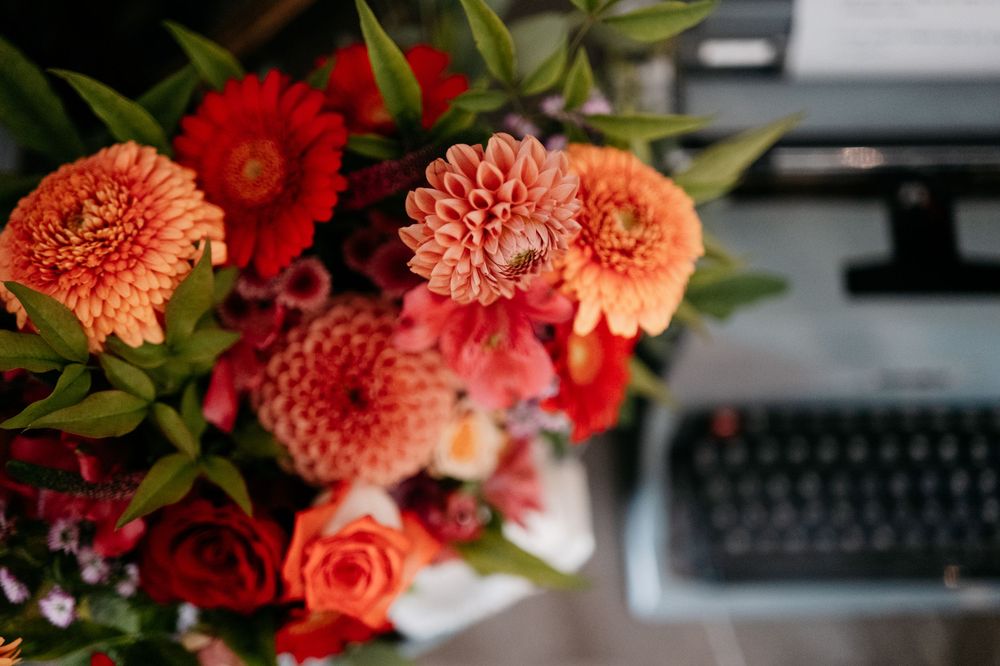 Vibrant coral and red dahlias with roses arranged next to a vintage typewriter.