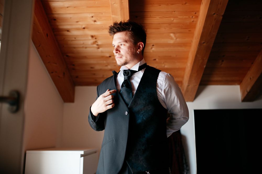 Groom adjusting tie in rustic wooden cabin with natural lighting.