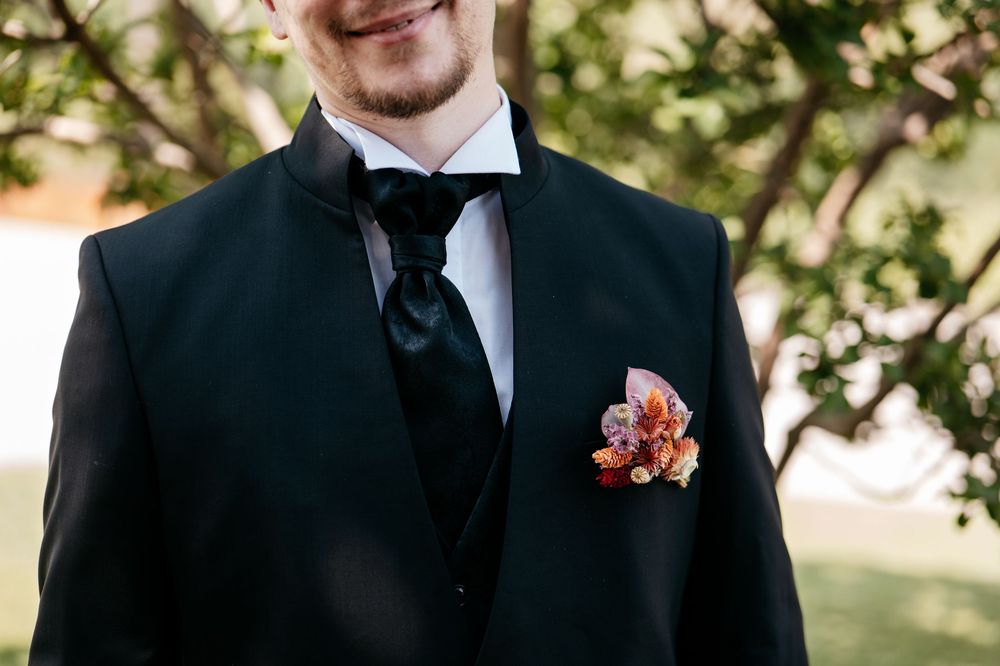 Close-up detail of a black tuxedo with coral boutonniere against a blurred natural background.