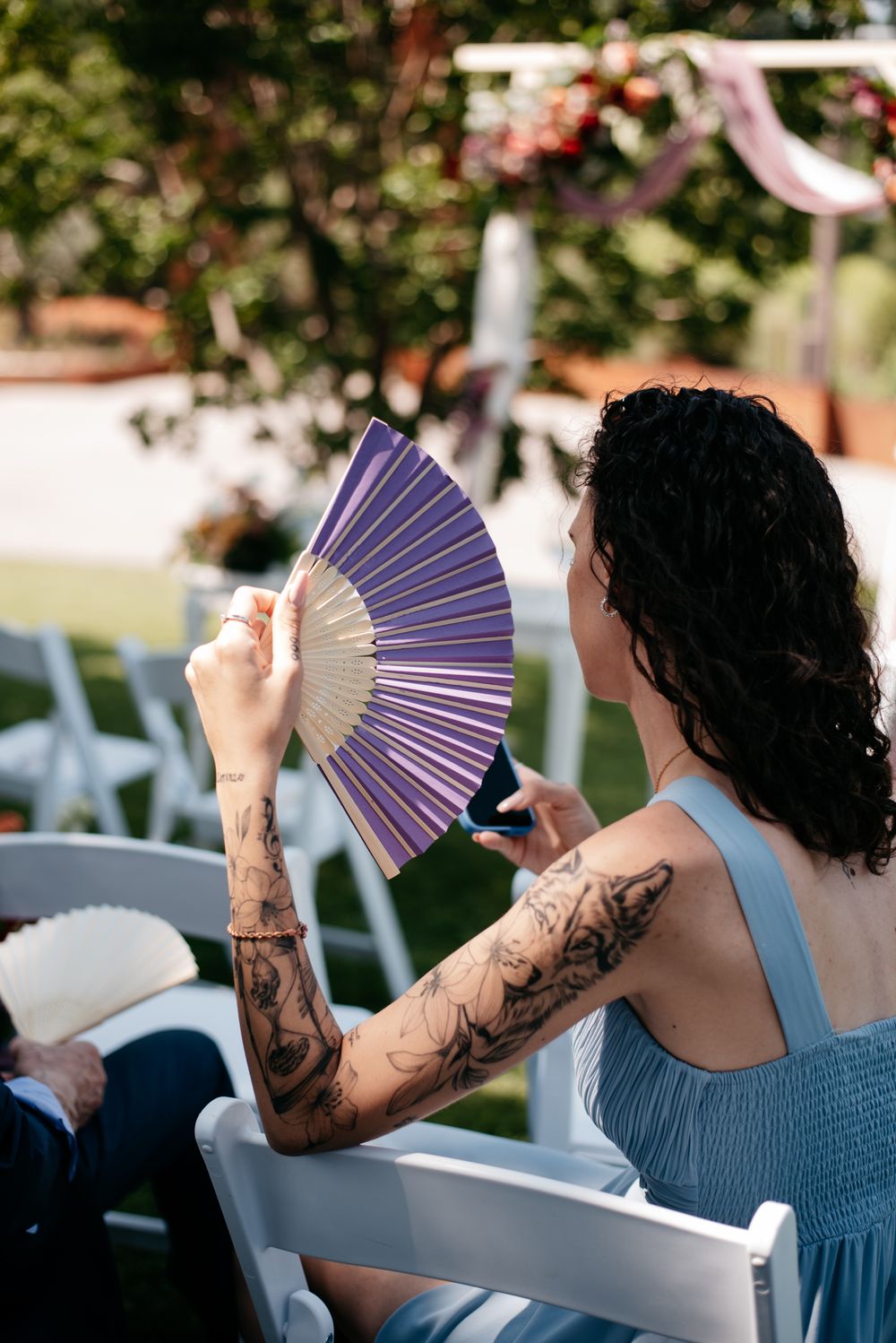 Person in blue dress using decorative purple and white hand fans at an outdoor event.