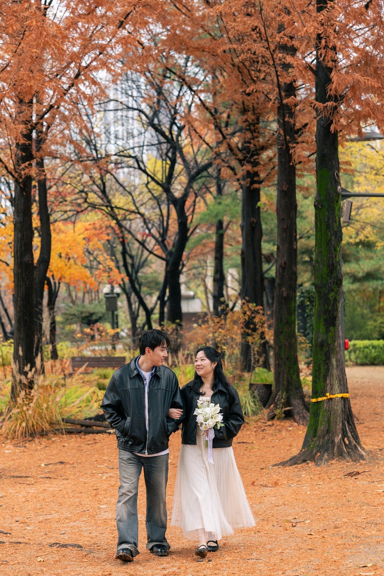 A couple walks together on a path covered in vibrant orange autumn leaves beneath tall trees with fall foliage.